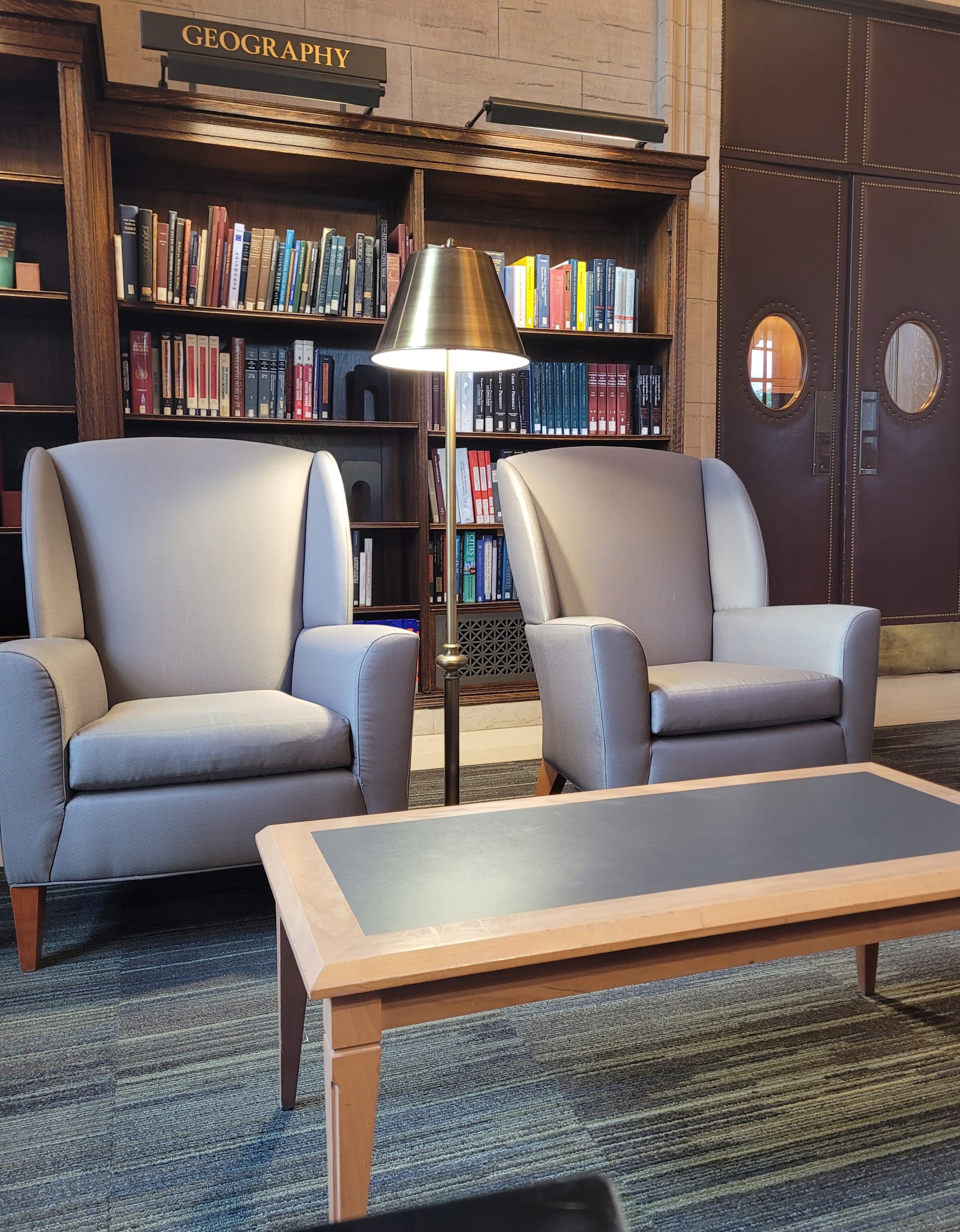 Two gray wingback chairs and a low rectangular table in front of a bookshelf labeled "Geography"